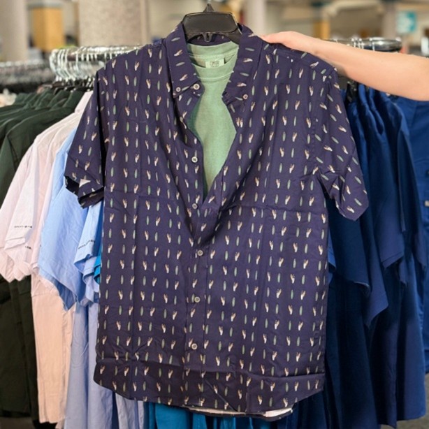 A patterned short-sleeve shirt featuring a navy background with white feather-like designs, held up by a person in a clothing store. A light green t-shirt is positioned underneath the shirt.