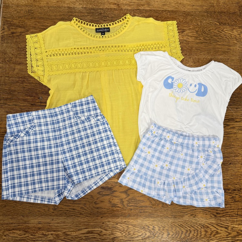 A yellow lace-accented blouse sits next to a white t-shirt with a blue and yellow graphic reading "Good things take time." Two pairs of shorts are also displayed: a blue and white plaid pair and a light blue pair decorated with daisies.